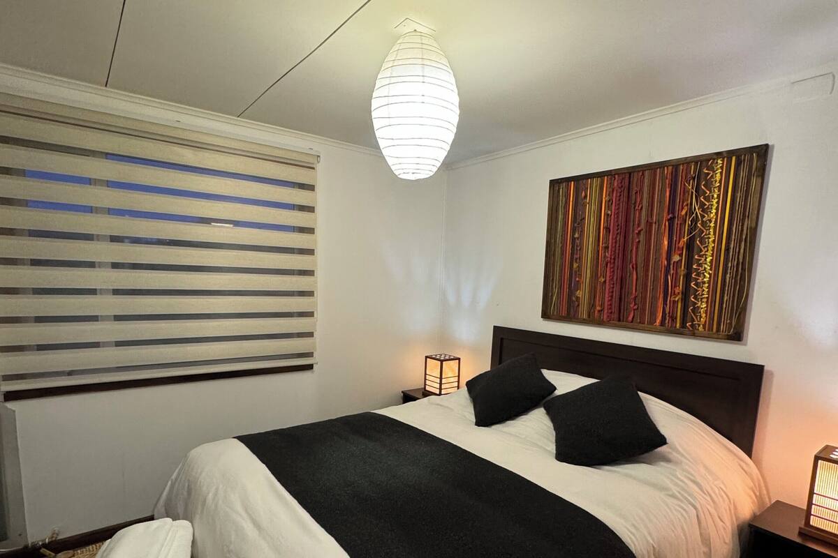 Una cama bien hecha con edredón blanco y negro, una lámpara de noche, una obra de arte en la pared y una ventana con persianas.