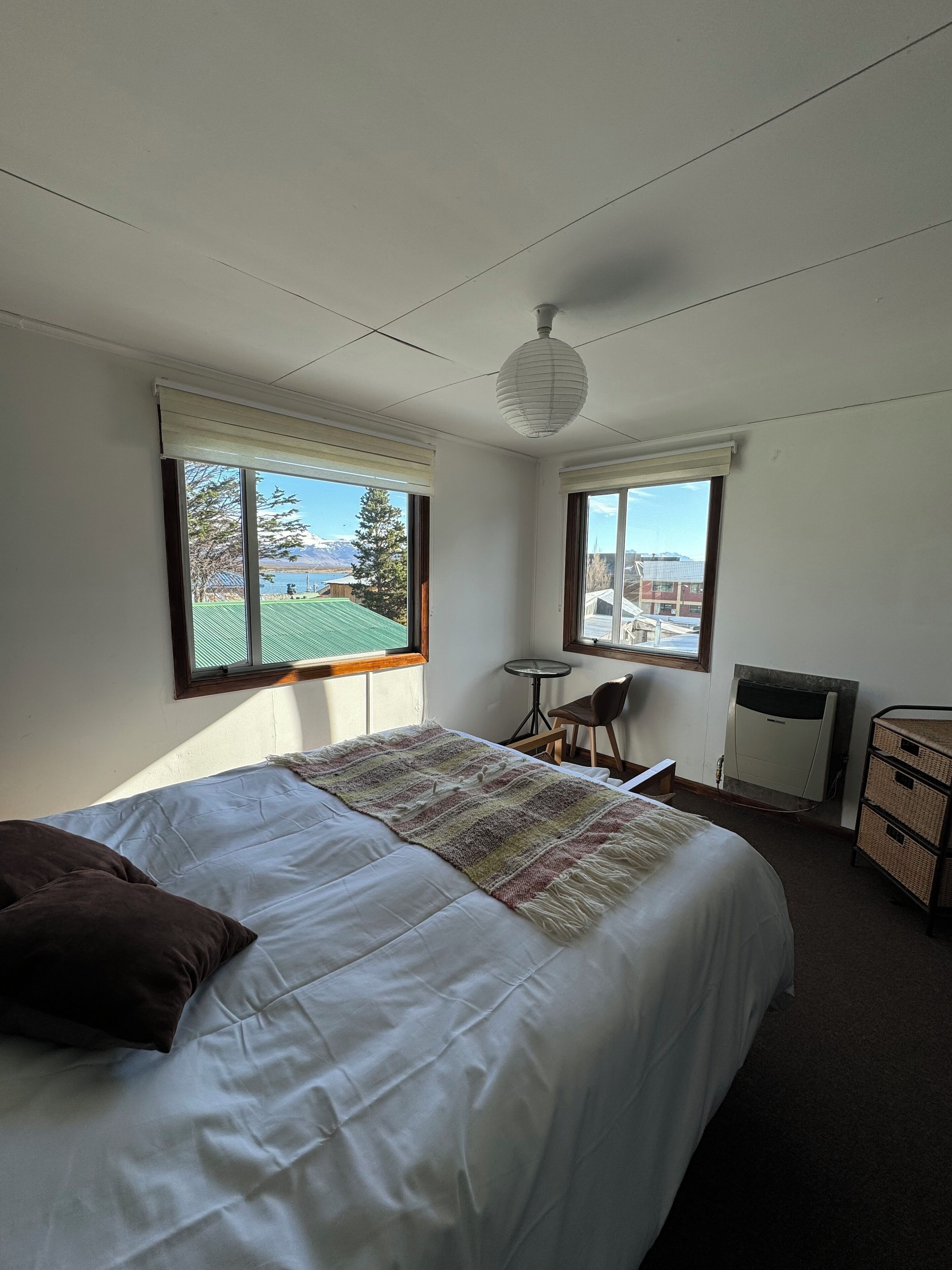 A bedroom with a bed, two windows, a heater, a small table, and a chair.