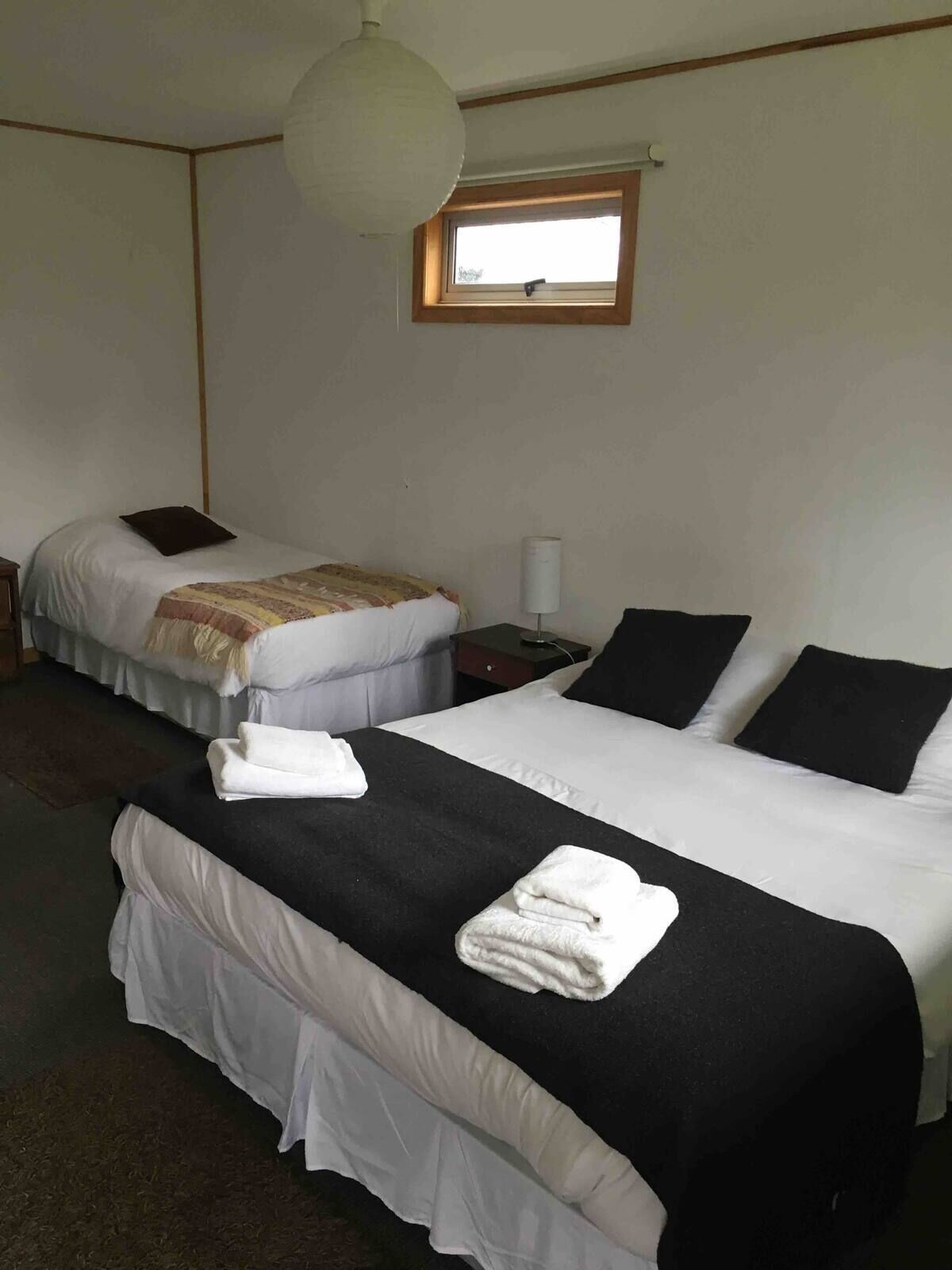 Een hotelkamer met twee eenpersoonsbedden, een klein raam, een nachtkastje met een lamp en een hanglamp.