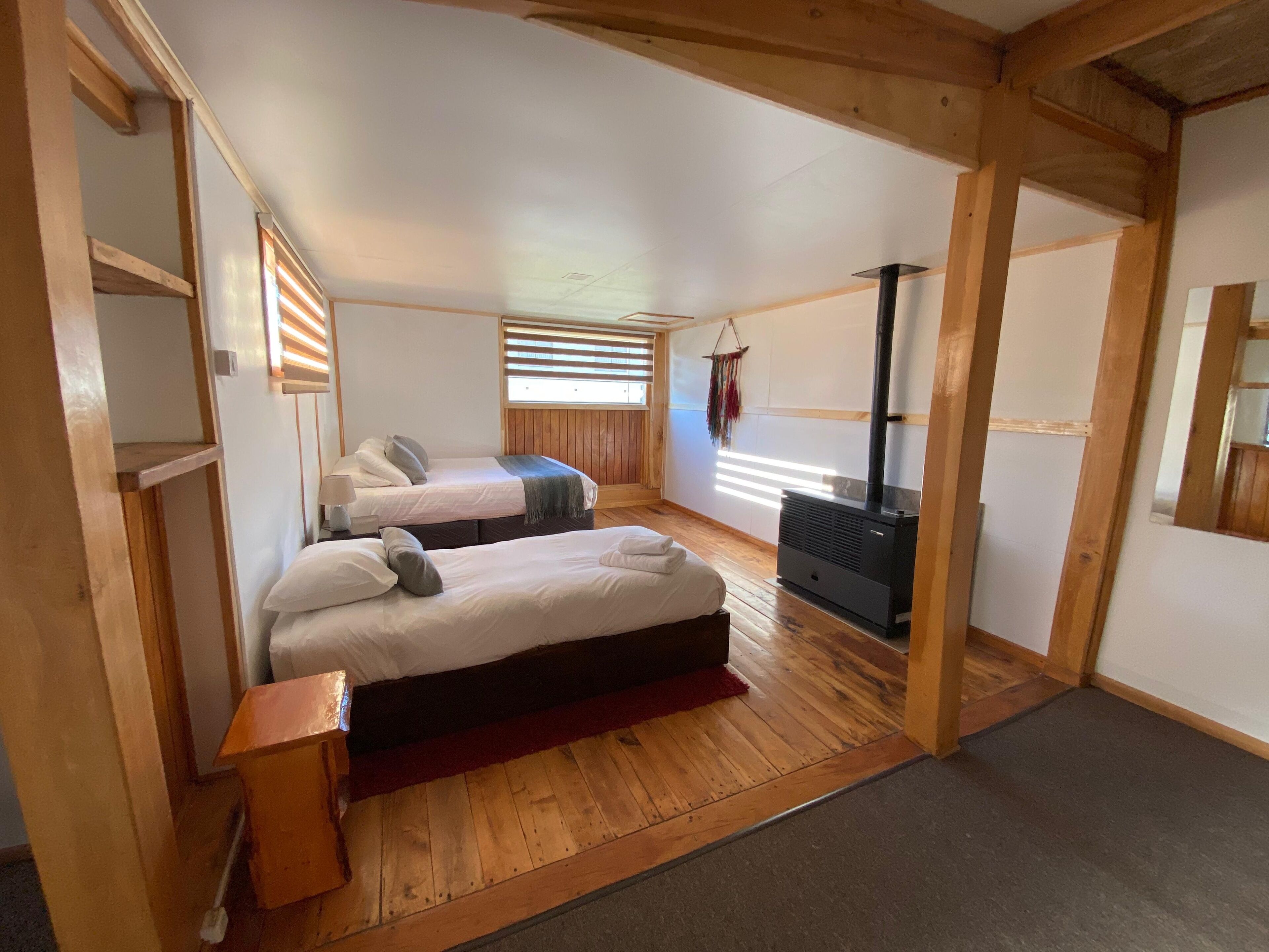 A cozy room with two beds, a wooden floor, a fireplace, and a window with blinds.