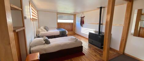 A cozy room with two beds, a wooden floor, a fireplace, and a window with blinds.