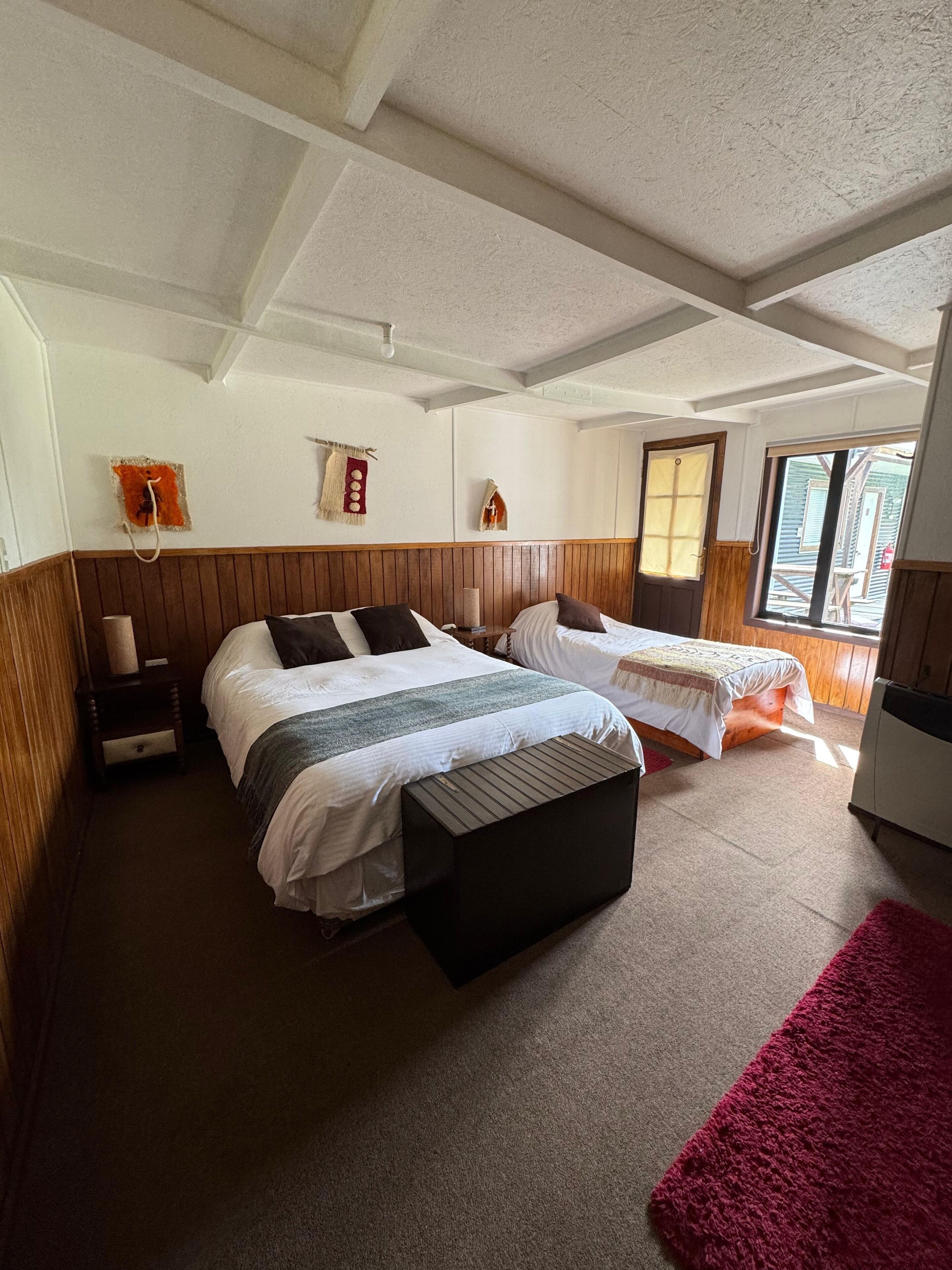 A bedroom with two beds, a wooden headboard, a nightstand, a red rug, and a window with a view of the outside.