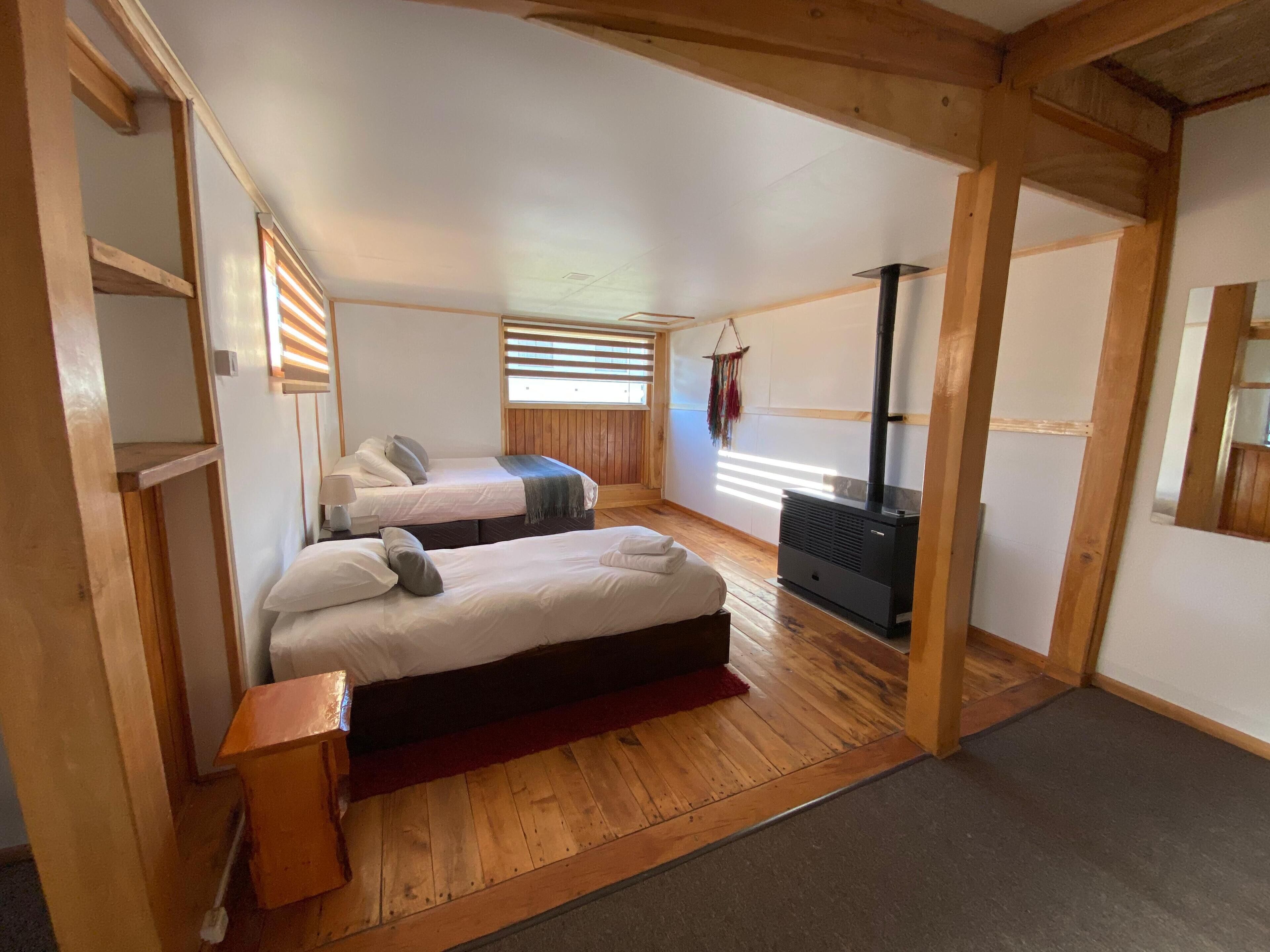 A cozy room with two beds, a fireplace, wooden beams, and a window with blinds.