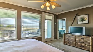 A hotel room with a bed, a television, a ceiling fan, and large windows with blinds. - Hole-in-One Vista! (Laguna Vista)