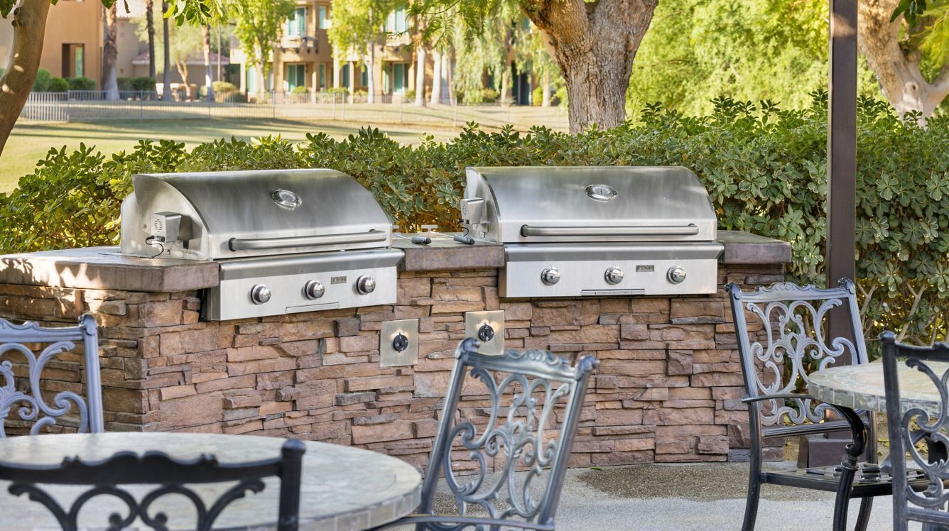 An outdoor patio with two stainless steel grills, stone wall, and wrought iron chairs.