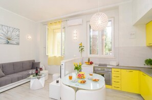 A modern living room with a grey sofa, a glass coffee table, and a dining area with a white table and yellow cabinets. - Cozy condo with AC, WiFi in charming Dubrovnik (Dubrovnik)