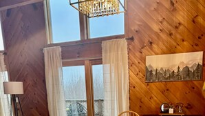 A wooden dining table with a reflective surface, a chandelier, and a view of a snowy landscape through a large window. - Cozy Chalet – Mtn View+ Hot Tub+ Hike+ Santa's Vil (Lancaster)
