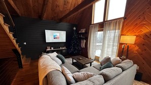 A cozy cabin interior with a wooden staircase, a gray sofa, a small table, and a Christmas tree. - Cozy Chalet – Mtn View+ Hot Tub+ Hike+ Santa's Vil (Lancaster)