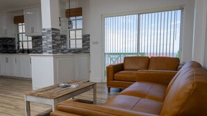 A modern living room with a brown leather sofa, a wooden coffee table, and a kitchen with white cabinets and a stone backsplash. - Sea and Summit Suite #1 (Castle Comfort)