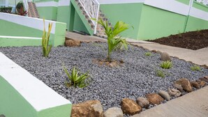 A landscaped garden area with a small palm tree, surrounded by gravel and rocks, and a staircase leading to a green building with white railings. - Sea and Summit Villa (Roseau)