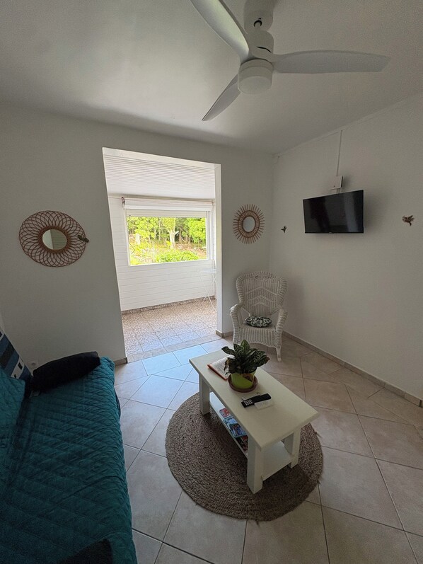 A living room with a ceiling fan, a television, a sofa, a chair, and a window with a view of greenery.