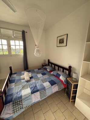 A bedroom with a bed, a bedside table, a nightstand, a hanging mosquito net, a window with curtains, and a framed picture on the wall.