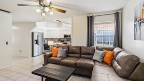A spacious living room with a brown leather sectional sofa, a wooden coffee table, and a ceiling fan. A kitchen is visible in the background with modern appliances. - 5/3 Vacation home with a large pool near the Disney attractions!! (Davenport)