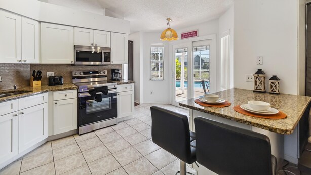 A modern kitchen with white cabinets, stainless steel appliances, and a granite countertop. There is a dining area with black chairs and a small table. - 5/3 Vacation home with a large pool near the Disney attractions!! (Davenport)
