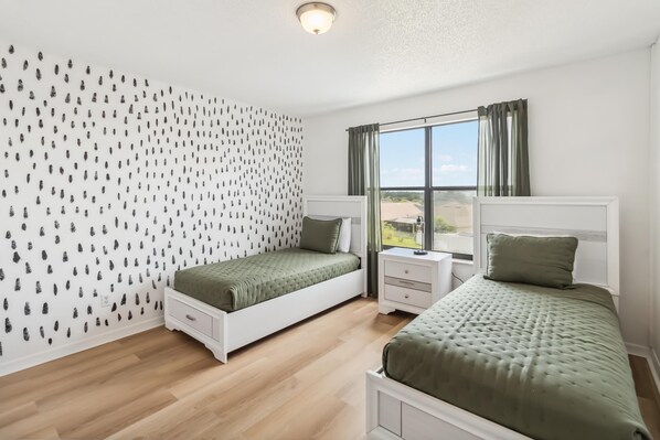 Une chambre avec deux lits simples, une fenêtre avec des rideaux, une table de chevet blanche et un mur orné d’un motif noir et blanc.