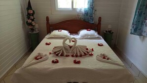 Una cama bien tendida con un arreglo de toallas en forma de corazón y pétalos de rosa, una ventana con cortinas, y una mesita de noche verde con un jarrón de flores.