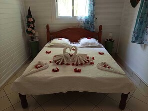 A neatly made bed with a heart-shaped towel arrangement and rose petals, a window with curtains, and a green side table with a vase of flowers.