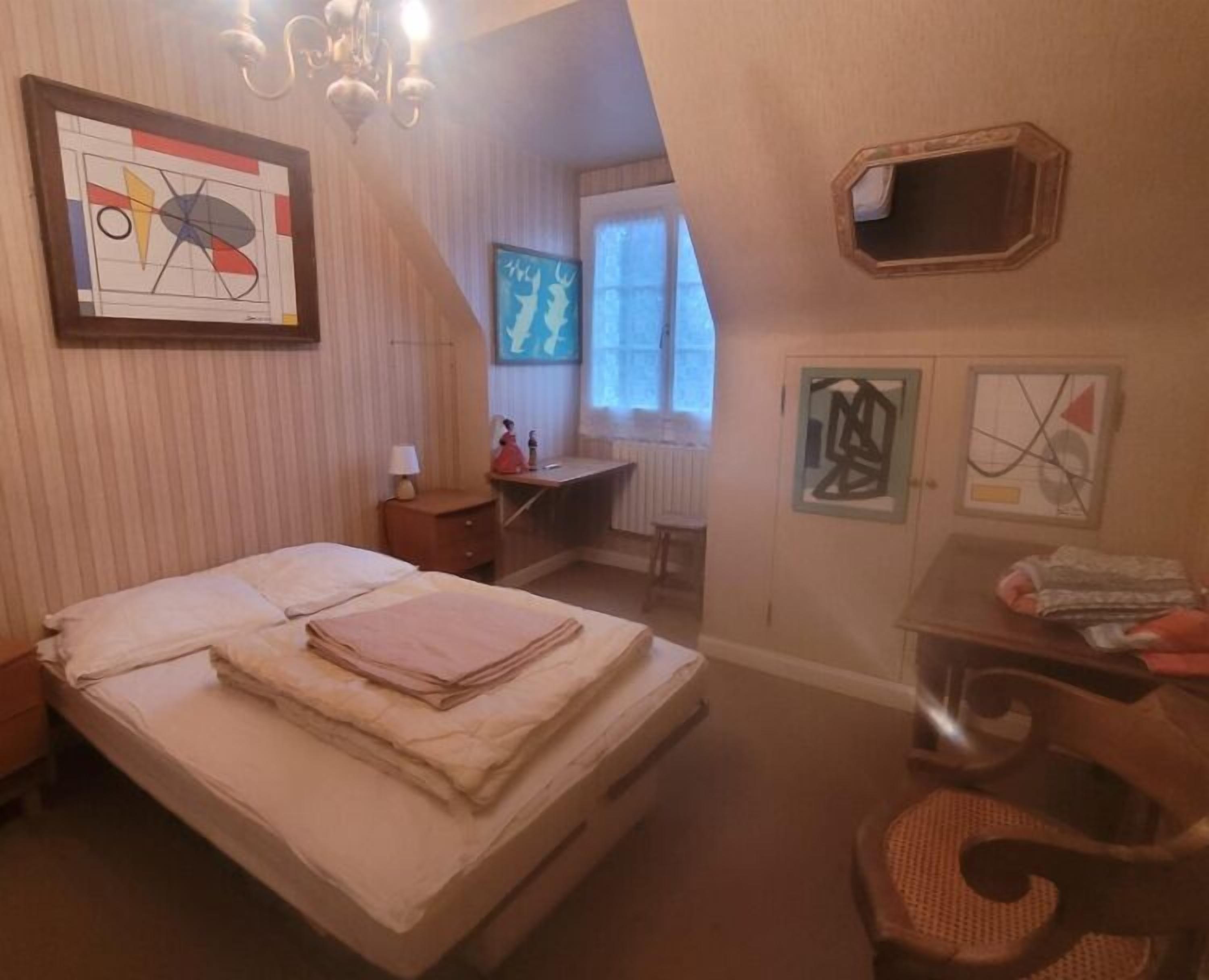 Une chambre à coucher avec un lit, un bureau et une chaise. Les murs sont ornés d’œuvres d’art encadrées et il y a une fenêtre avec des stores.