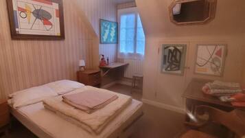 Une chambre à coucher avec un lit, un bureau et une chaise. Les murs sont ornés d’œuvres d’art encadrées et il y a une fenêtre avec des stores.