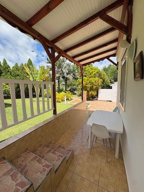 Un patio cubierto con mesa y sillas, un banco y vistas a un jardín con diversas plantas y árboles.