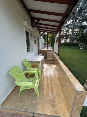 Un patio techado con una mesa y sillas, piso de baldosas y vista a un jardín con palmeras.