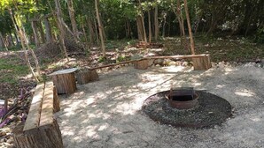 A forested outdoor area with a circular fire pit surrounded by gravel, wooden benches, and a rustic wooden bridge.