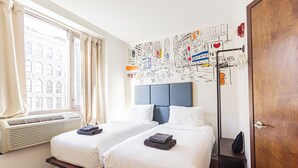 A hotel room with two beds, a large window with curtains, a wall with a cityscape mural, and a wooden door.