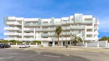 Un edificio blanco de varios pisos con balcones, una palmera frente a él y automóviles estacionados en la calle.