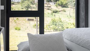 Quarto com uma janela grande, cama com roupa de cama branca e vista para uma paisagem rural.