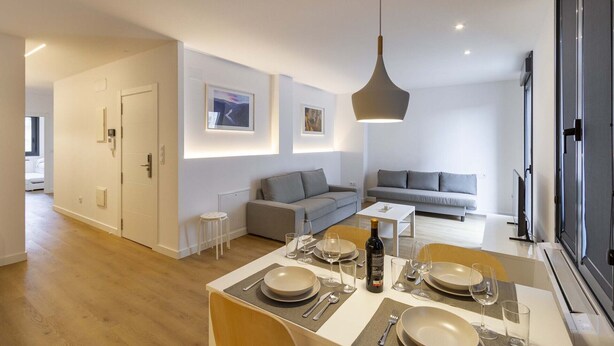 Uma sala de estar moderna com área de jantar, sofás cinzentos e uma grande janela.