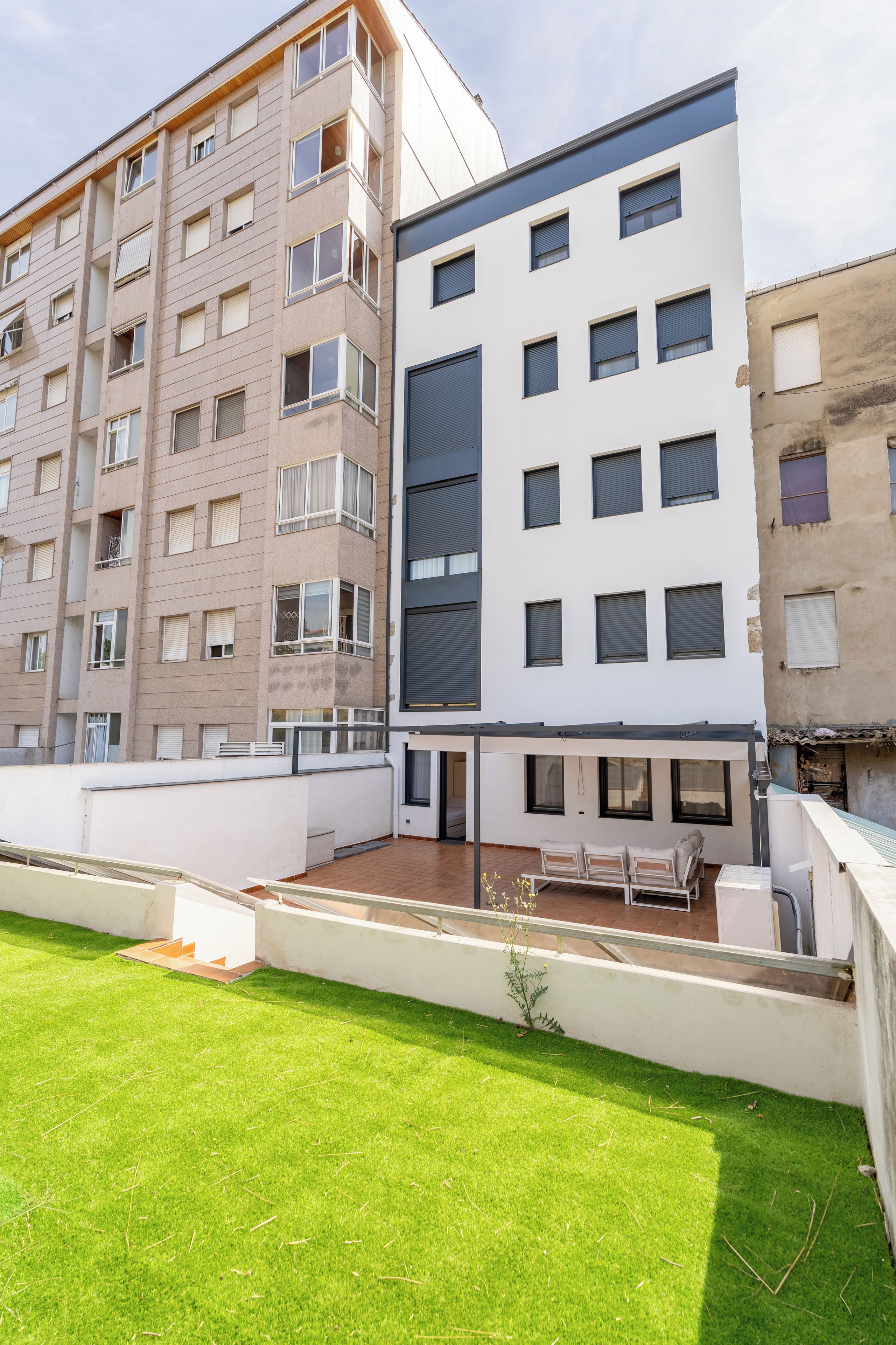 Un edificio residencial de varios pisos con balcones, una terraza en la azotea plana y un pequeño jardín.