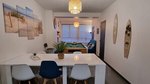 A modern kitchen with a dining area, featuring a pool table, a hanging light fixture, and surfboards on the wall.