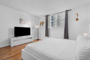 Una habitación de hotel moderna con una cama grande, un televisor de pantalla plana, una mesita de noche, una lámpara y una ventana con cortinas.