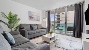 A modern living room with a grey sofa, a coffee table, and a large window with a view of a balcony and buildings.
