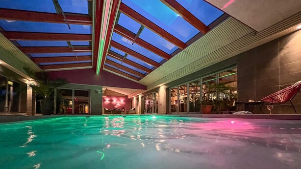 An indoor swimming pool with a glass ceiling, a bar area, and a lounge chair.