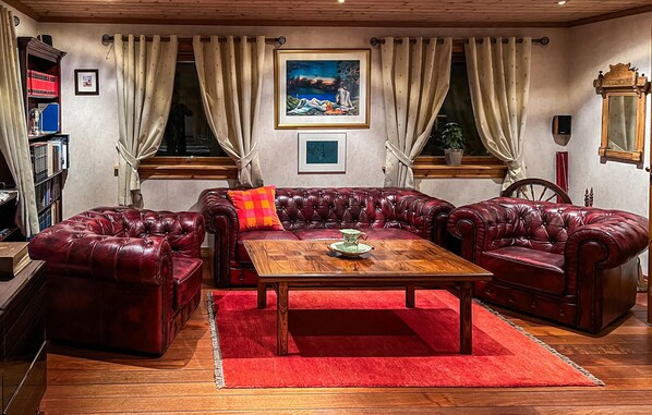 Una sala de estar acogedora con un sofá de cuero rojo, una mesa de centro de madera y una alfombra roja. Hay dos ventanas con cortinas, un cuadro en la pared y una estantería con libros y un objeto rojo.