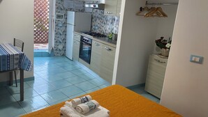A compact kitchen with a refrigerator, stove, and oven. There is a dining table with chairs and a small table with a basket of flowers. - Ponzamania Siren (Le Forna)