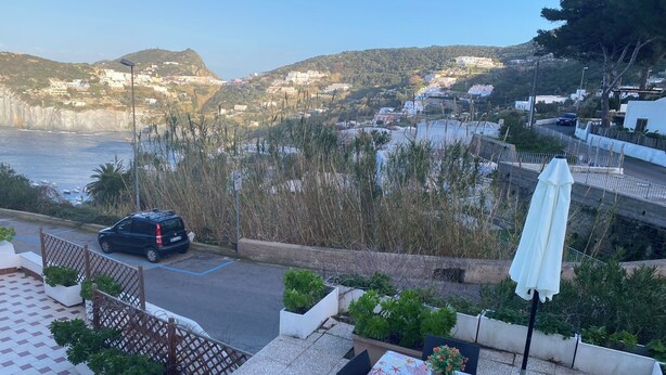 A terrace with a table set for dining, overlooking a coastal town with buildings on a hillside and a parked car. - Ponzamania Siren (Le Forna)