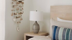 Un lit bien fait, avec une couette blanche, une tête de lit en bois, une lampe de chevet et un carillon éolien fixé au mur.