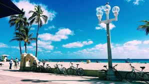 A beachside promenade with bicycles parked along the path, palm trees, and a clear blue sky.