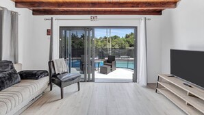 A modern living room with a sofa, a chair, a TV, and a sliding glass door leading to a pool area.