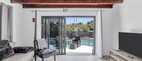 A modern living room with a sofa, a chair, a TV, and a sliding glass door leading to a pool area.