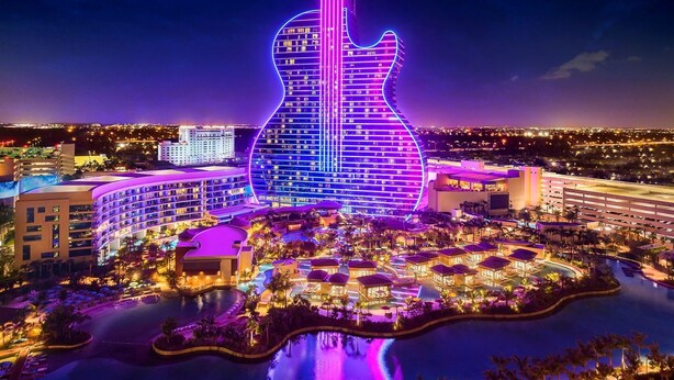 A hotel with a guitar-shaped building illuminated in purple and blue lights, surrounded by a water body and palm trees.