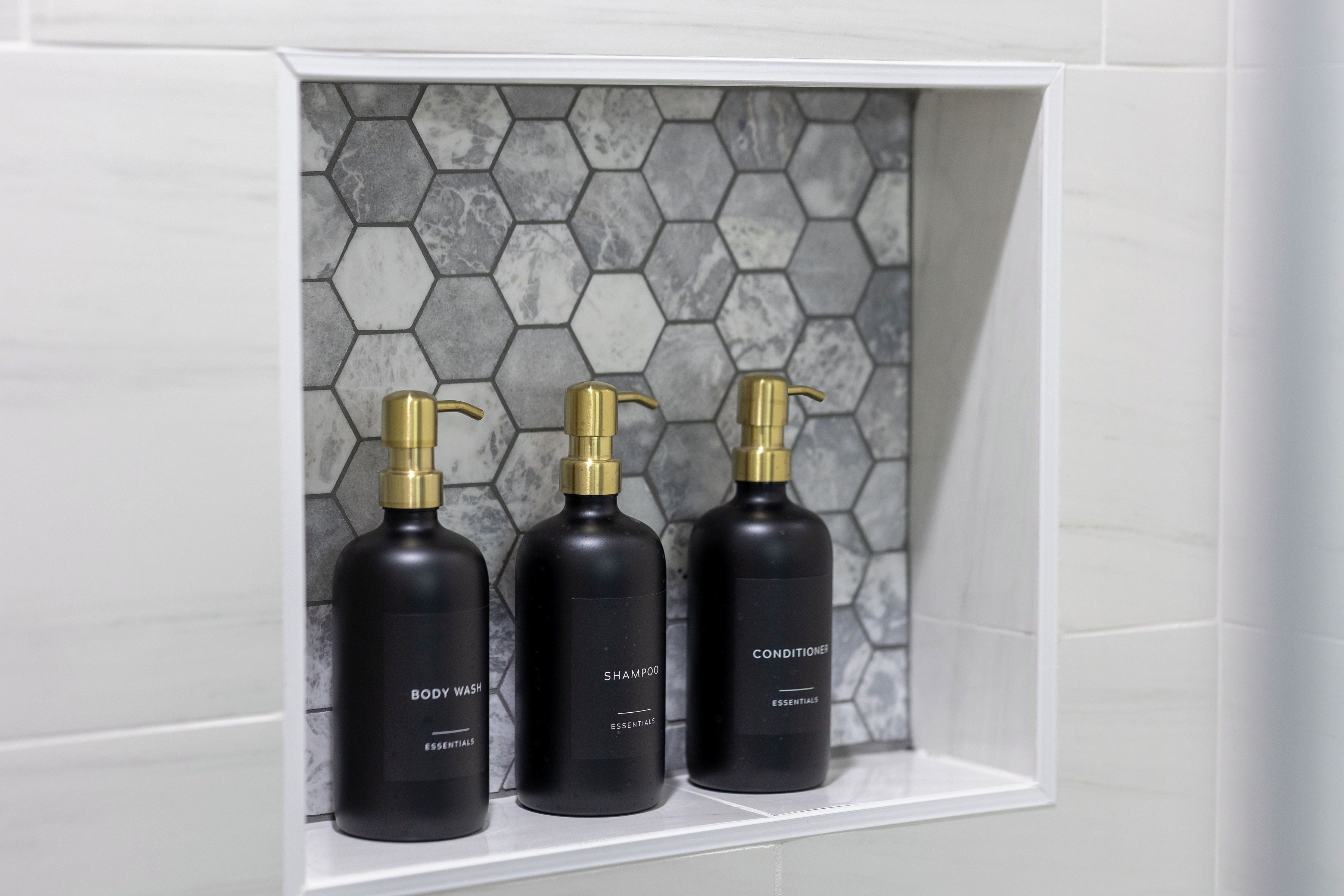 Three black bottles with gold pumps on a white shelf against a hexagonal tile wall.
