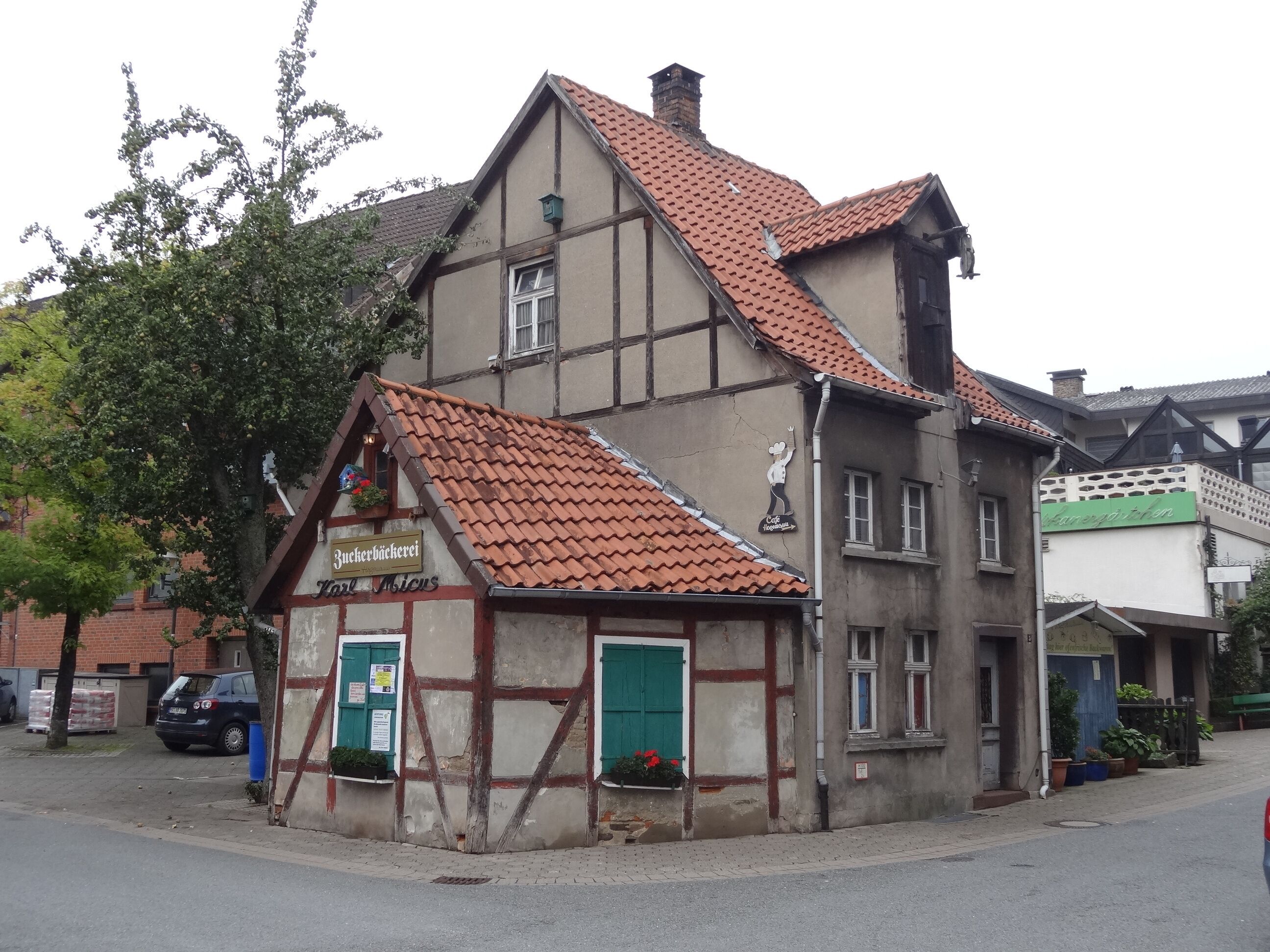 Un bâtiment traditionnel à colombages, surmonté d’un toit de tuiles rouges, avec une petite dépendance dotée d’un volet vert et une enseigne visible indiquant la présence d’un café.