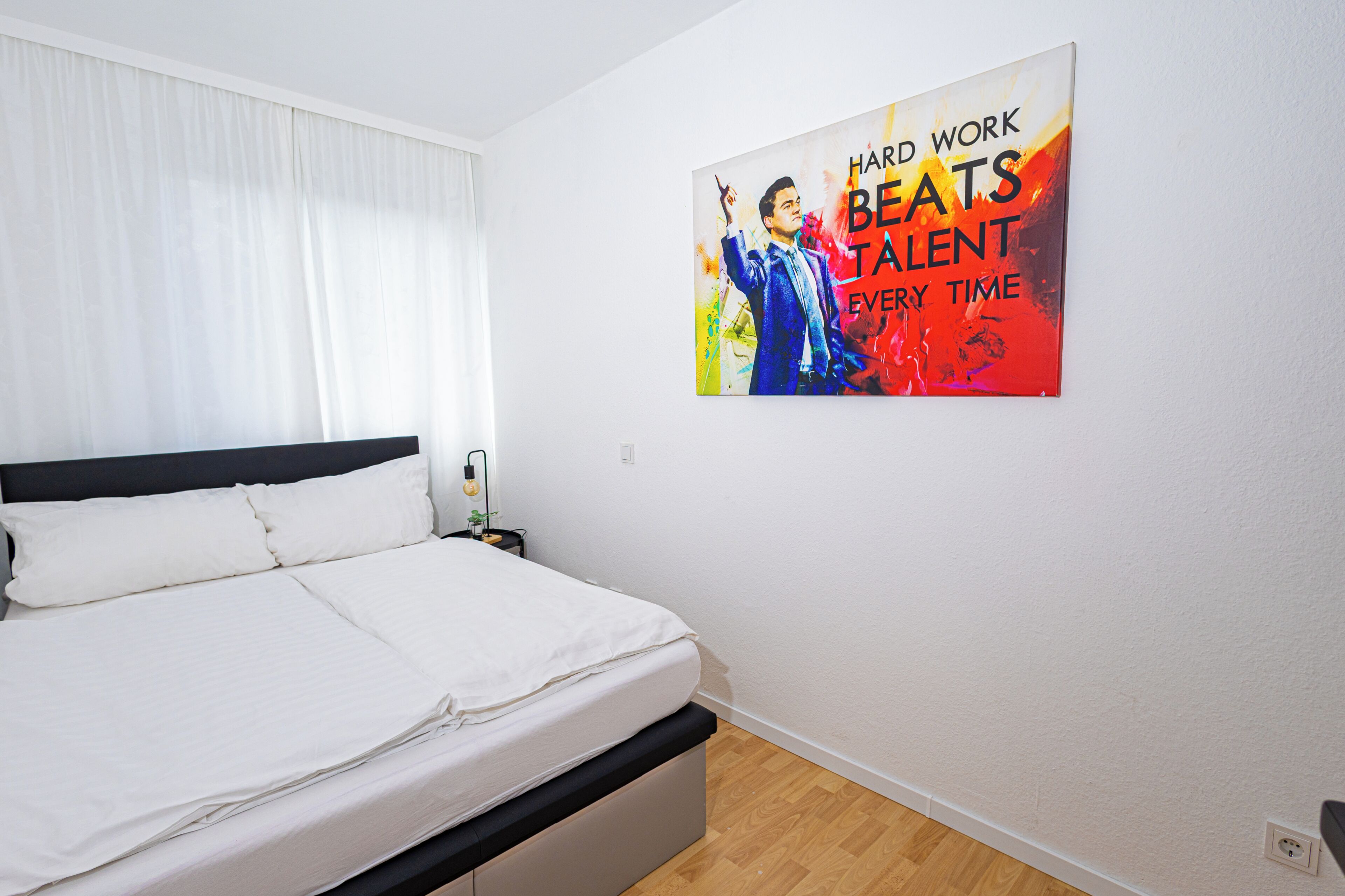 Een slaapkamer met een bed, een nachtkastje, een wanddecoratie en een raam met gordijnen.