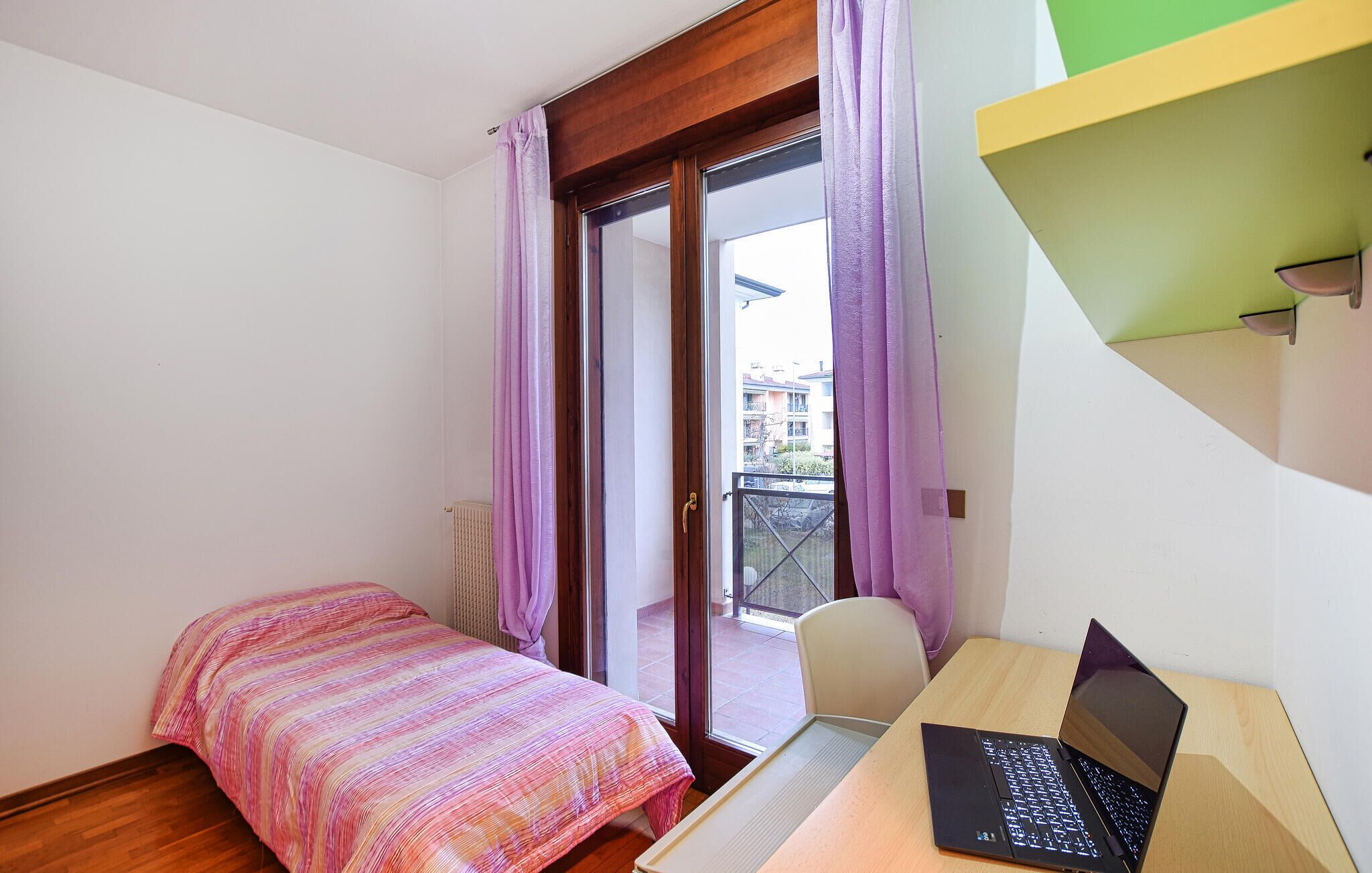 A small bedroom with a bed, a desk with a laptop, and a sliding glass door leading to a balcony.