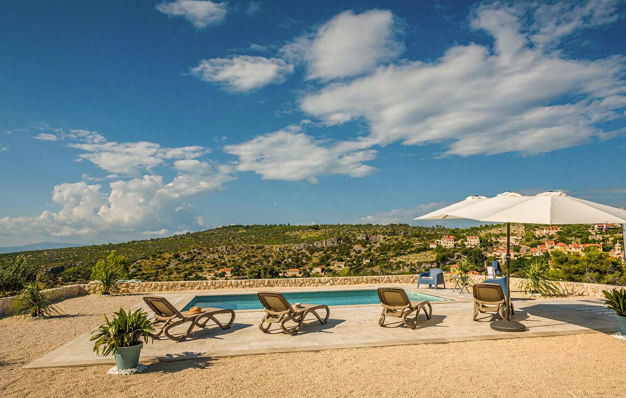 A poolside area with lounge chairs and a large umbrella, overlooking a hillside with buildings.