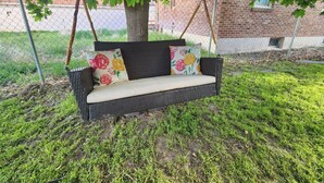 Una silla colgante de mimbre con cojines estampados, ubicada debajo de un árbol, y un edificio de ladrillo al fondo.