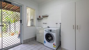 A compact laundry room with a washing machine, a sink, and a window with blinds. - Agnes Break - Sauna, Surfboards, Walk To Beach (Agnes Water)